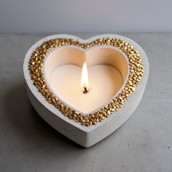 Heart-shaped concrete candle holder with a lit white candle and gold beads around the rim, resting on a gray surface, conveying a romantic/cozy mood.
