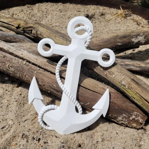 White decorative anchor with a rope wrapped around it, resting on sand with driftwood nearby.
