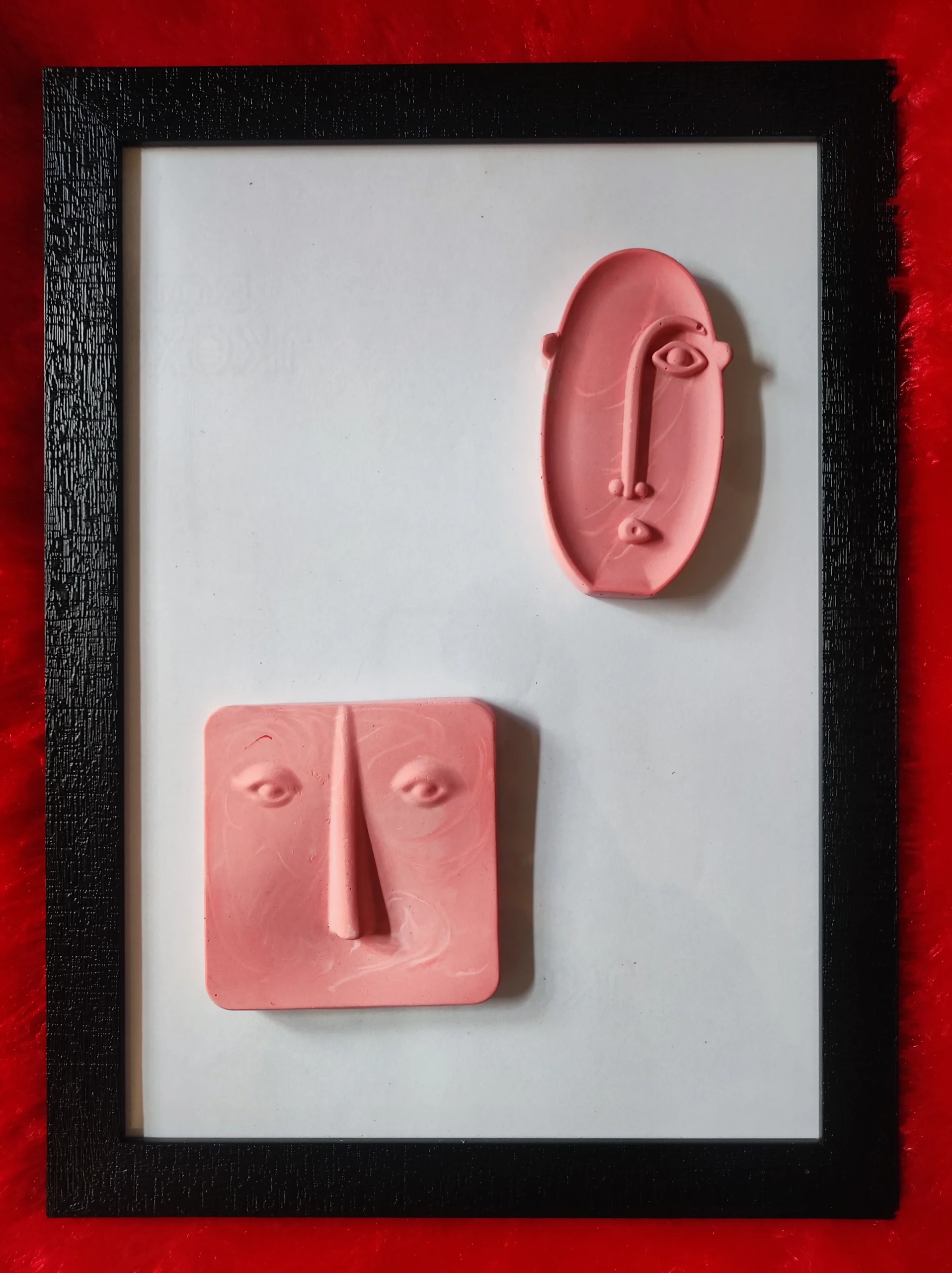 Pink clay sculpture of a stylized face with an elongated nose, set inside a black picture frame on a white mat with a red background.