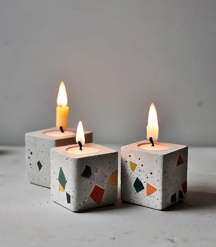 Three square terrazzo candles with colorful geometric flecks, lit and burning on a light gray surface.