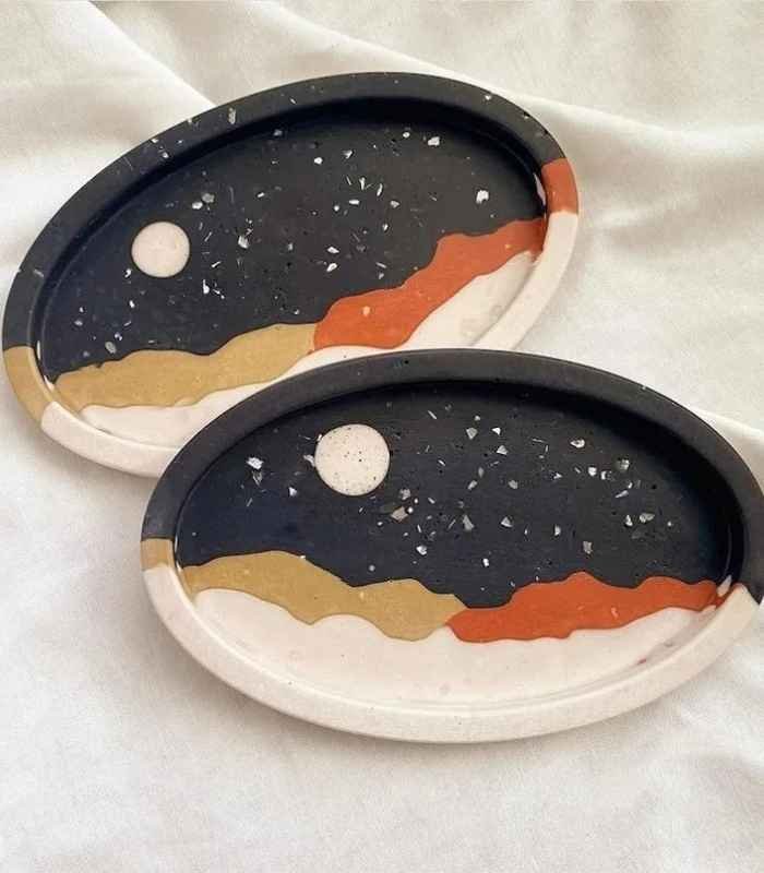 Two oval decorative trays with a night-sky design: dark background, white moon, and layered gold, beige, orange, and white waves.