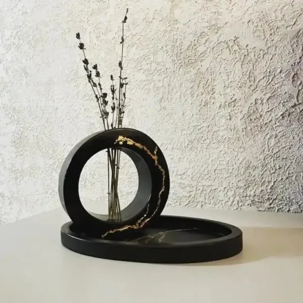 Black circular sculpture with a hollow ring and a vase of dried stems, resting on a round base against a textured wall.