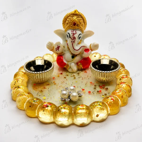 Small white Ganesha figurine with a gold crown sits at the center of a circular ring made of gold beads on a glittery base. Two decorative bowls flank the statue, and scattered confetti and pearl earrings lie in front.
