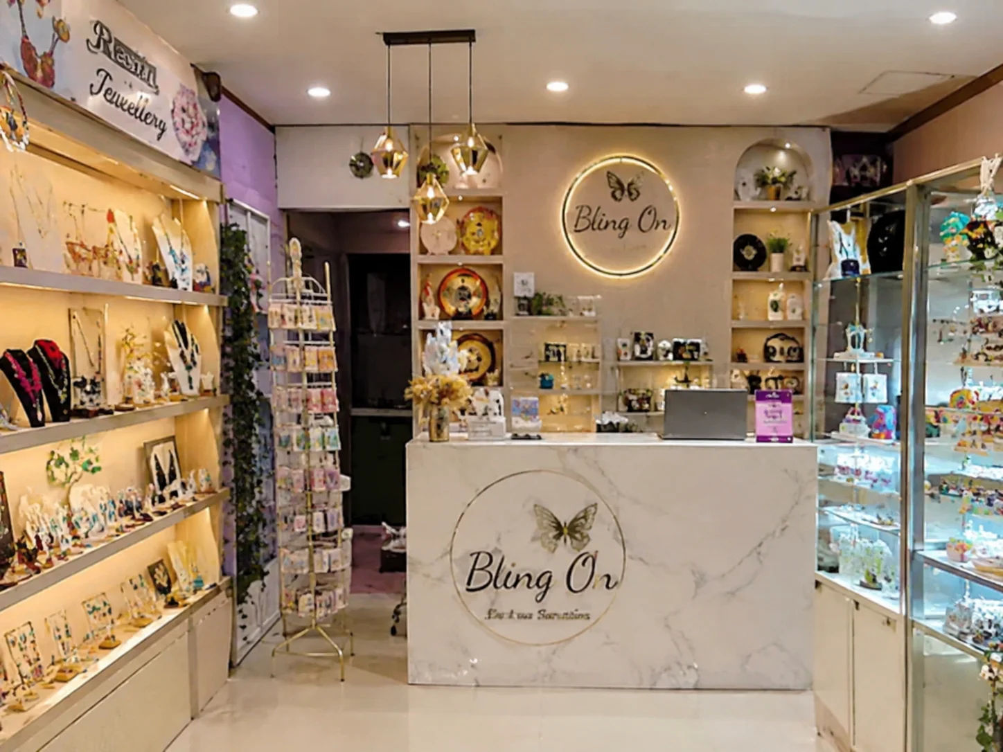 Inside a bright jewelry shop called Bling On, with a marble-front counter featuring a butterfly logo and shelves of necklaces and decor around the room.